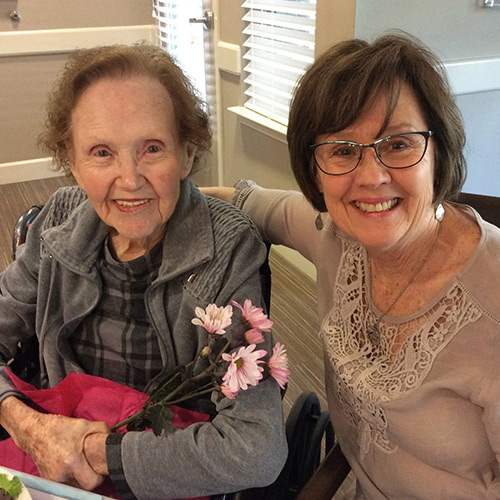 Two elderly women smiling indoors, with one holding pink flowers. Both are wearing glasses and casual clothing. The background features a window with blinds and a table with a red cloth.