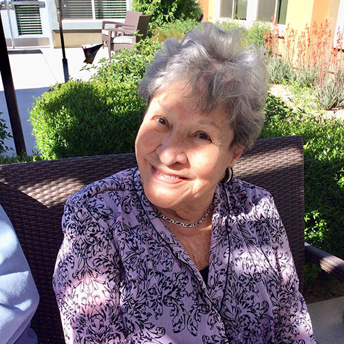 A smiling elderly woman with curly gray hair sits outdoors on a brown chair, wearing a patterned purple top. Sunlight creates a dappled effect on her face and clothing. She appears relaxed and cheerful in a garden setting.