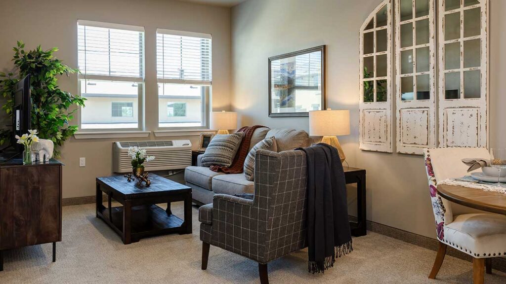 A cozy living room with a gray sofa, patterned armchair, and wooden coffee table. Two table lamps are lit, and a vintage window frame decorates one wall. A small dining area with a round table is visible, and two large windows let in natural light.