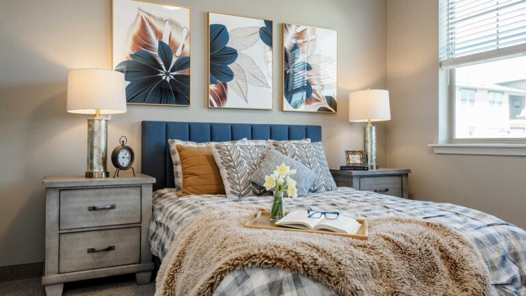 A cozy bedroom with a bed featuring a blue upholstered headboard, floral art above, and two gray nightstands with lamps. The bed is adorned with patterned pillows and a fur throw. A tray with a book, glasses, and flowers sits on the bed.