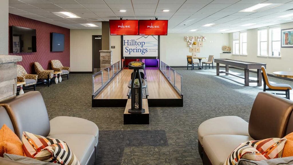 A recreational room with a mini bowling lane and seating area. The walls are decorated, and there's a sign that reads "Hilltop Springs." Natural light comes in through large windows on the right.