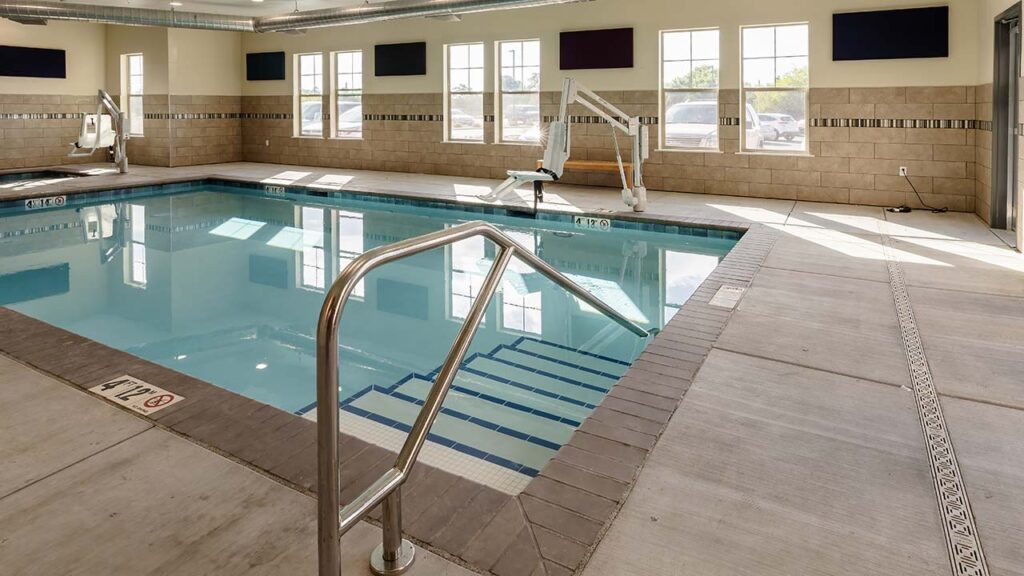 Indoor swimming pool with clear water, surrounded by beige tiles and a metal handrail leading down steps into the pool. Large windows allow natural light to illuminate the area. Pool signs indicate a depth of 4 feet.