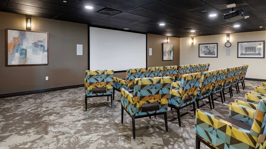 A small theater room with geometric-patterned chairs arranged in rows facing a large white screen on a beige wall. Abstract art and framed photos decorate the walls. The ceiling is black with recessed lighting. Carpet features a neutral pattern.