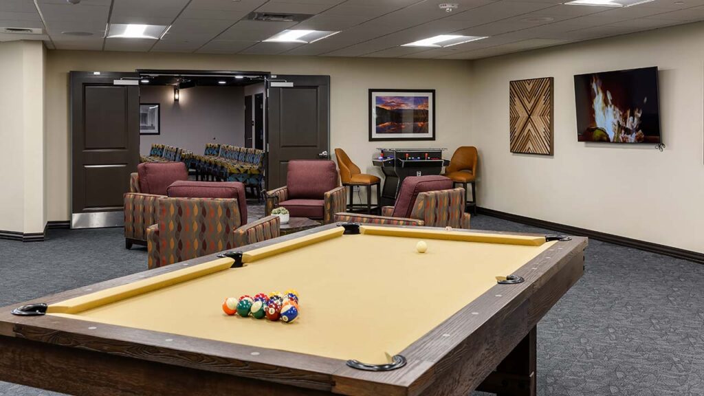 A recreation room with a pool table in the foreground. The table has a set of pool balls ready for play. In the background, there are several cushioned chairs, a flat-screen TV on the wall, and framed artwork.