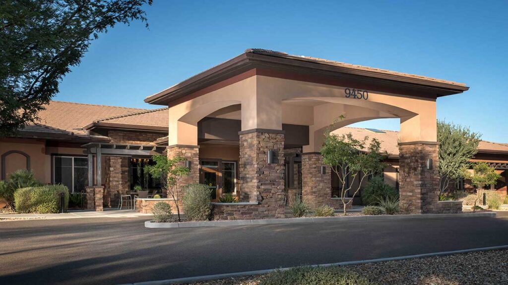 A modern building with stone pillars and a large overhang at the entrance. The exterior features earthy tones and desert landscaping, including shrubs and small trees. The building number 9450 is displayed above the entrance.