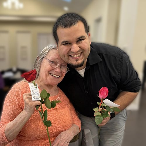 An elderly woman in an orange top and a smiling man in a black shirt pose together, each holding a red rose. They are indoors, with a blurred background.