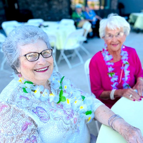 Two elderly women smiling and seated at an outdoor event. They are wearing floral leis and glasses. Tables with pale yellow tablecloths and more people in the background are visible. The scene is bright and cheerful.