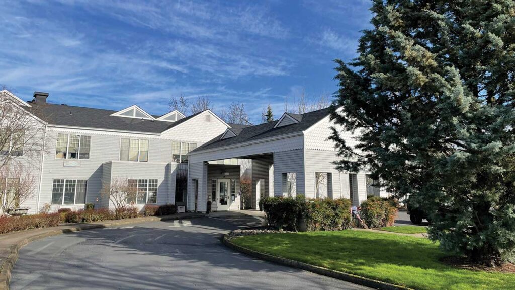 A two-story building with light gray siding, featuring large windows and a covered entrance. The foreground shows a curved driveway, green grass, and a tall evergreen tree. The sky is blue with light clouds.