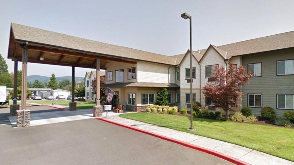Exterior view of a two-story building with a covered entrance and well-maintained landscaping. The structure features neutral-colored siding and a sloped roof, with a driveway and a small lawn area. The setting is bright and clear.