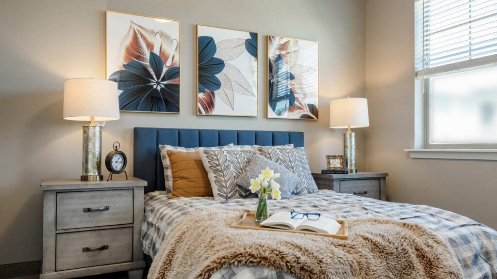 A cozy bedroom with a bed featuring beige and gray bedding, accented with a faux fur throw. Two bedside tables with lamps flank the bed. Three floral artworks hang above the dark blue headboard. A tray with a book and flowers rests on the bed.