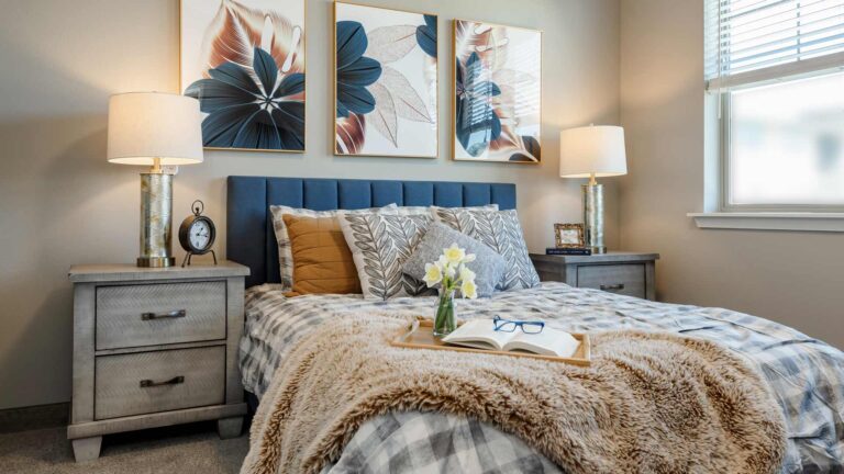 A cozy bedroom with a plaid bedspread, a blue upholstered headboard, and decorative pillows. A tray with flowers, an open book, and glasses sits on the bed. Two bedside tables with lamps and abstract floral artwork adorn the wall.