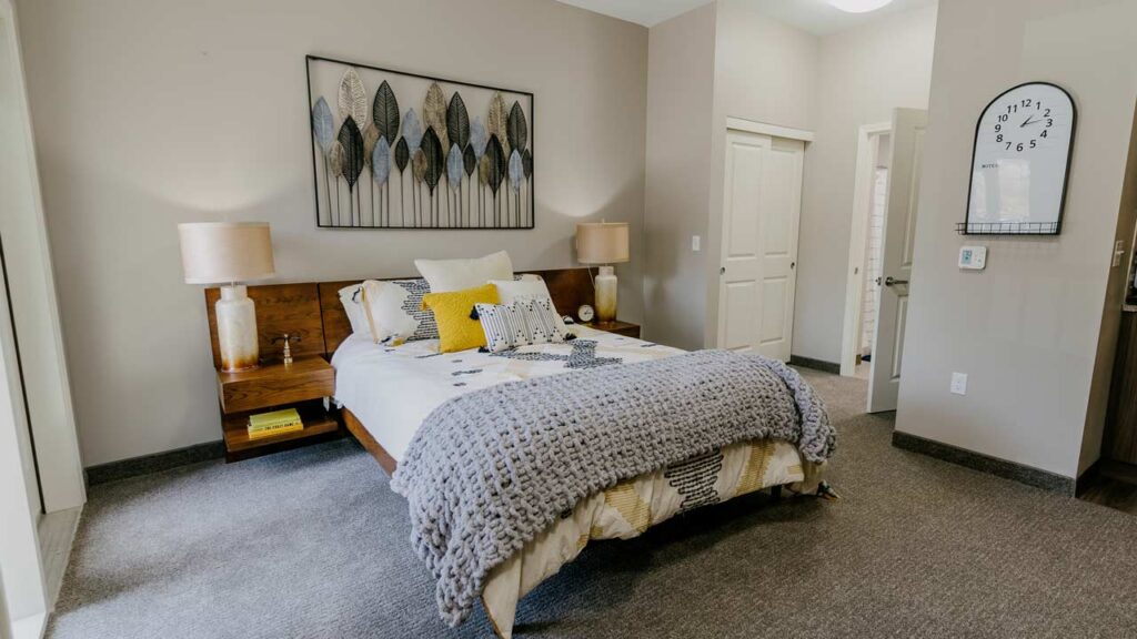 A cozy bedroom features a wooden bed with gray and yellow bedding, a knitted throw blanket, and decorative pillows. Two wooden nightstands with lamps flank the bed. Wall art with leaf designs hangs above the bed, and a round clock adorns the wall.