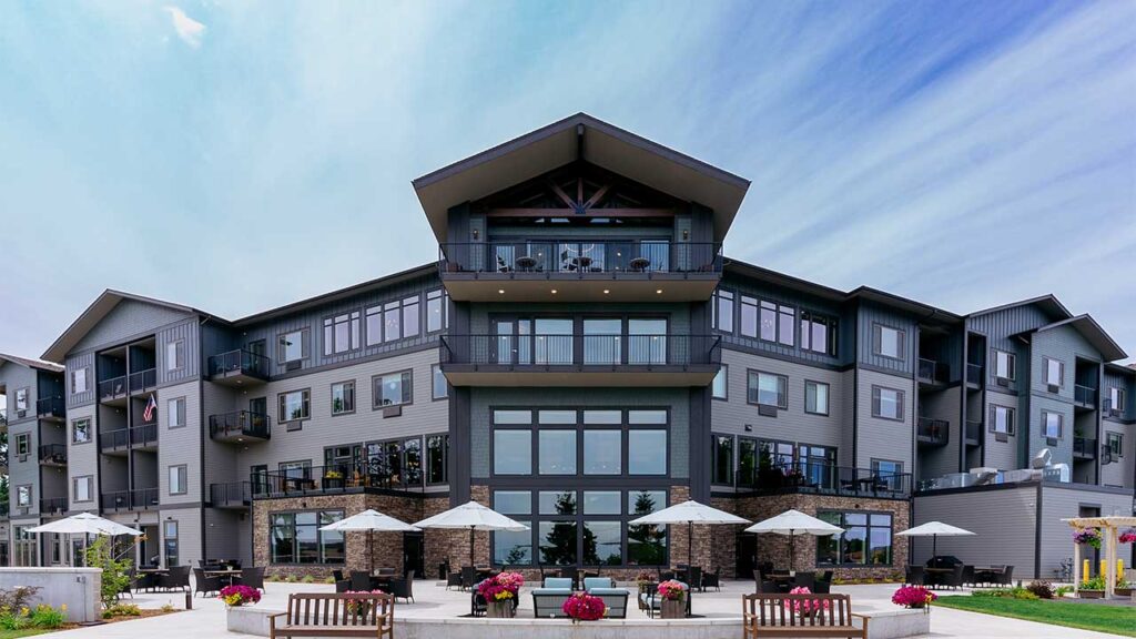 Modern three-story apartment building with large windows, balconies, and a peaked roof. The foreground features a patio area with umbrellas, tables, benches, and flower pots. The sky is partly cloudy.