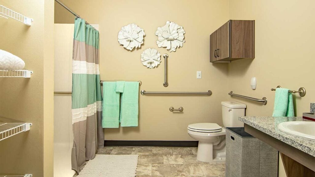 A bathroom featuring accessibility aids. It has a walk-in shower with a striped curtain, grab bars, and a toilet. Green towels hang on the wall, with decorative white flowers above. There's a sink with a granite countertop on the right.