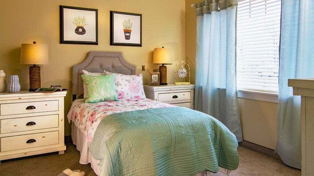 A cozy bedroom features a twin bed with floral bedding and a green quilt. The room has two white nightstands with lamps and decorative items. Two framed botanical prints hang on the wall above the headboard. Blue curtains cover the window.