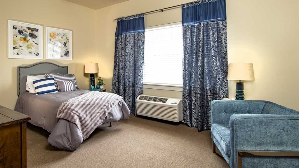 A cozy bedroom featuring a single bed with gray and striped bedding. Two floral paintings hang above the bed. Blue patterned curtains frame a window, and a blue armchair is placed beside the bed. Two lamps flank the bed on wooden end tables.