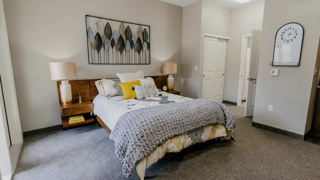 Cozy bedroom with a wooden bed, gray knitted blanket, and decorative pillows. Twin lamps on side tables flank the bed. Wall art features stylized leaves above the headboard. A round white clock is mounted on a beige wall. Doorway leads to another room.