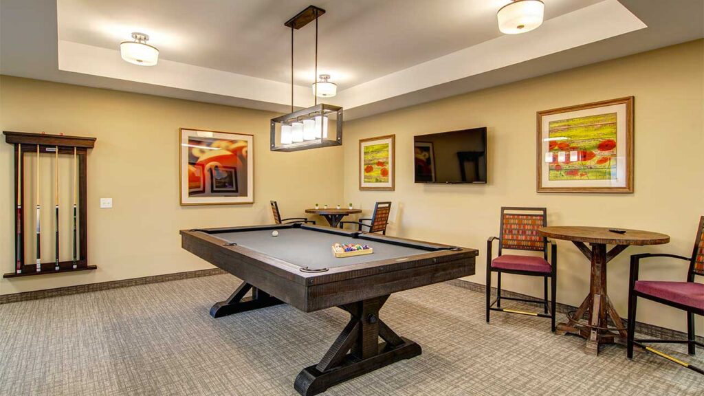 A modern game room with a billiards table in the center, pool cues on the wall, two chairs at a small round table, and a flat-screen TV mounted above. The room is warmly lit, with two framed artworks on the walls.