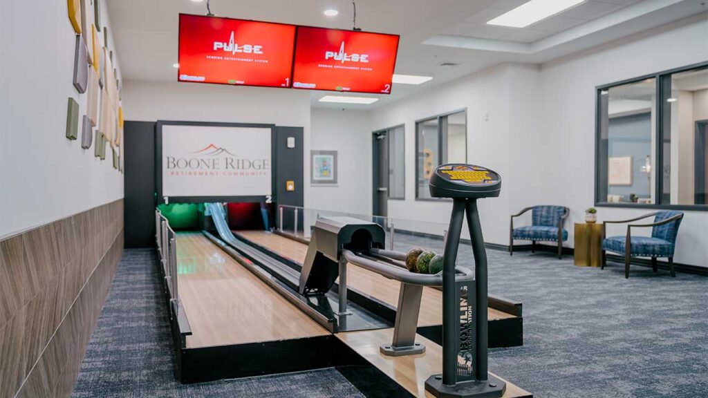 An indoor mini-bowling alley with a wooden lane, digital scoring screens, and a rack with bowling balls. The setting is cozy with blue chairs, wall art, and the sign "Boone Ridge Retirement Community" on the back wall.