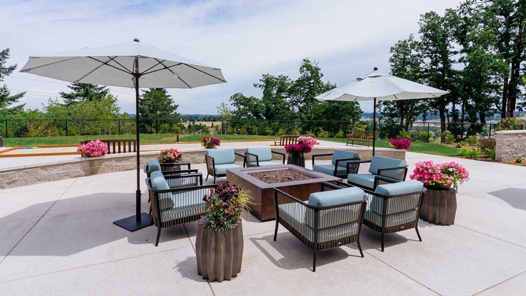 A patio with cushioned chairs surrounding a modern fire pit table. Two large umbrellas provide shade. Several planters with vibrant pink flowers enhance the outdoor setting. Trees and a scenic view are visible in the background.