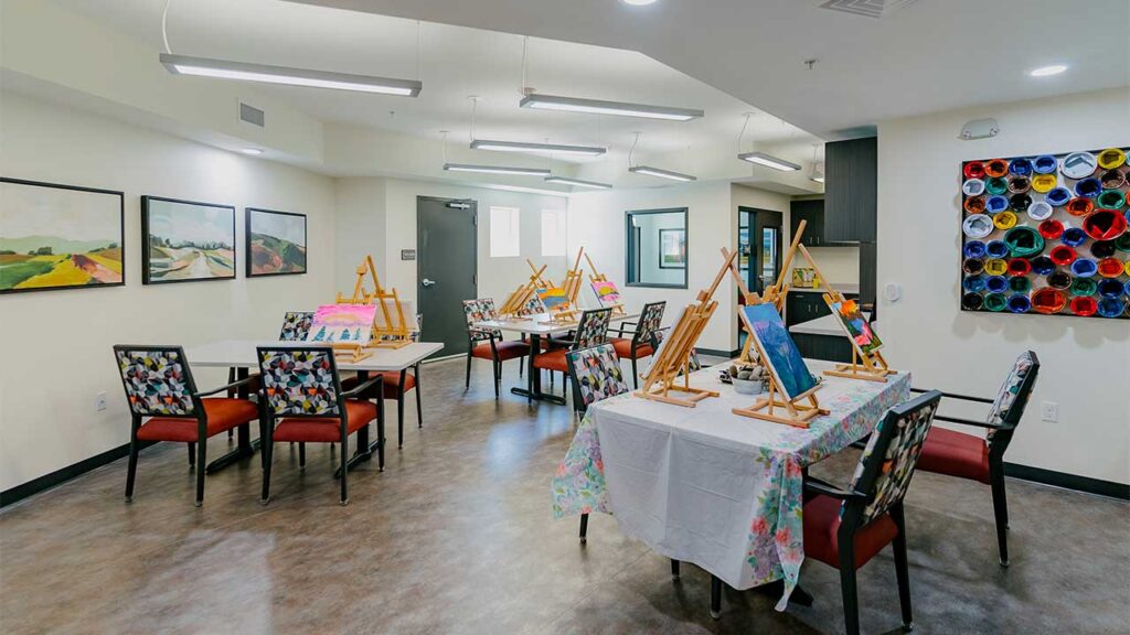 A well-lit art room with tables set up for painting. Easels hold canvases in progress, surrounded by chairs with floral cushions. The walls display landscape paintings, and shelves filled with colorful rolls are visible.