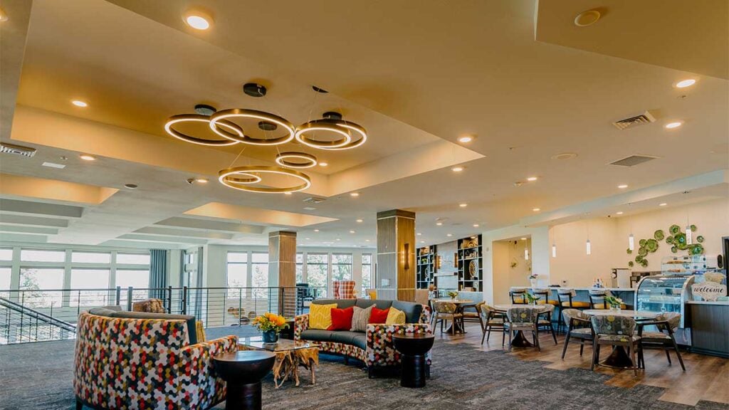 A modern lounge area with stylish circular light fixtures, colorful sofas, and wooden tables. The room features large windows, a cozy seating arrangement, and a small café on the right with tables and chairs, creating a warm, inviting atmosphere.