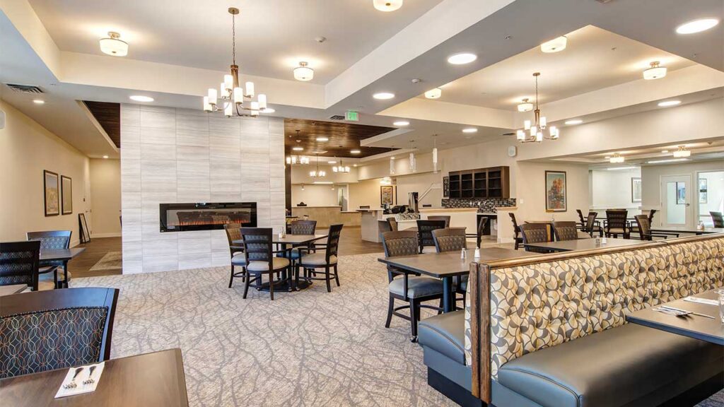 A spacious, modern restaurant dining area with a high ceiling and various seating options, including chairs and patterned booths. Elegant lighting fixtures hang from the ceiling, and there's a fireplace on the wall near the center.