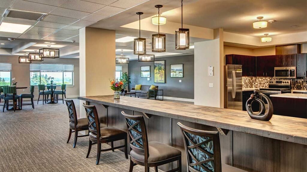 A modern kitchen and dining area featuring a long wooden counter with chairs, pendant lights hanging above, a stainless steel refrigerator, a cozy seating area with a sofa and paintings, and a dining table with chairs near large windows.