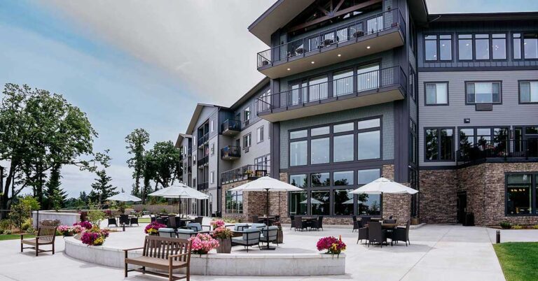 A modern multi-story building with large windows and balconies. The outdoor area features tables with umbrellas, benches, and potted flowers on a spacious patio. Surrounded by trees under a partly cloudy sky.