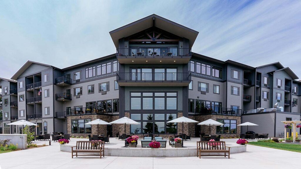Modern three-story apartment building with large windows and balconies. The foreground features a spacious patio area with benches, planters, and umbrella-covered tables. The sky is clear with a few wispy clouds.