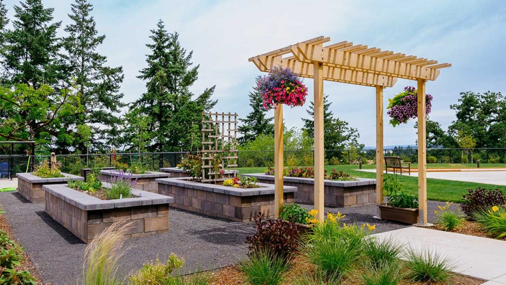 A garden with raised stone beds is adorned with a wooden pergola and hanging flower baskets. Lush greenery and tall trees provide a serene backdrop under a blue sky. A paved walkway runs alongside the garden.