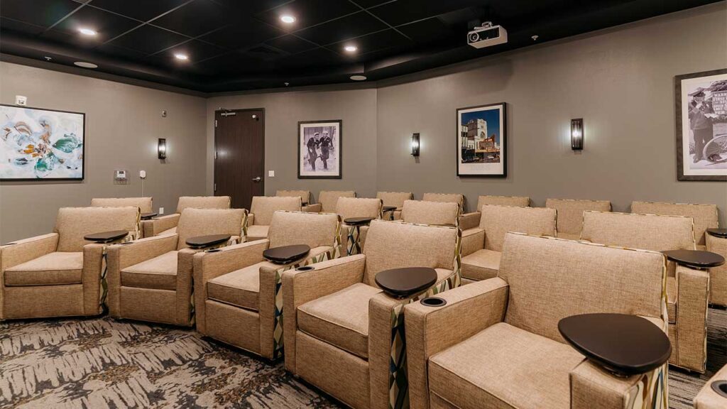 The image depicts a small, modern movie theater with rows of beige recliner seats, each equipped with a small table. The walls are adorned with framed artwork and dim lighting creates a cozy atmosphere.