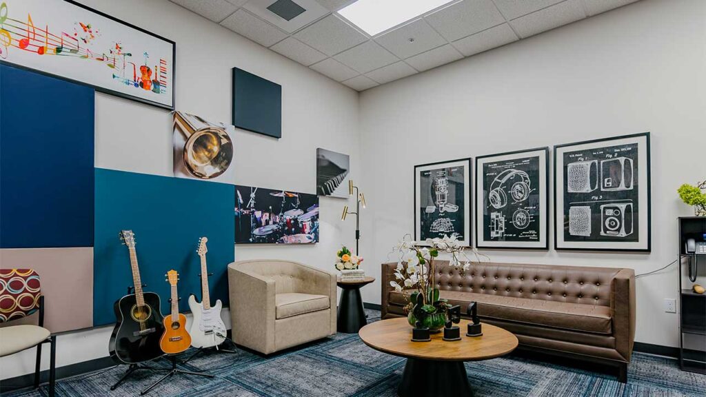 A cozy music-themed room with a beige armchair, brown leather couch, and round wooden table decorated with plants. Guitars and a ukulele rest on stands. The walls feature colorful art and instrument-themed prints. A coffee machine is in the corner.