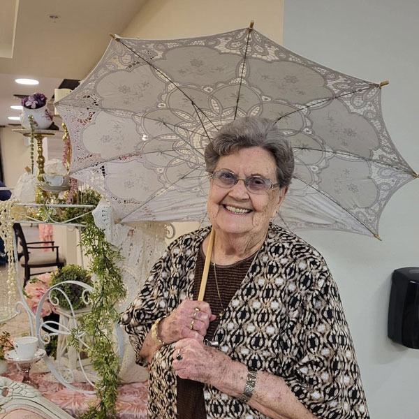 Elderly woman smiling broadly, holding a decorative lace parasol. She wears glasses and a patterned outfit, standing in a room with vintage decor, including plants and floral arrangements.