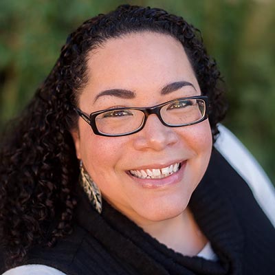 Smiling person with curly hair and glasses, wearing a black top and decorative earrings, with a blurred green background.