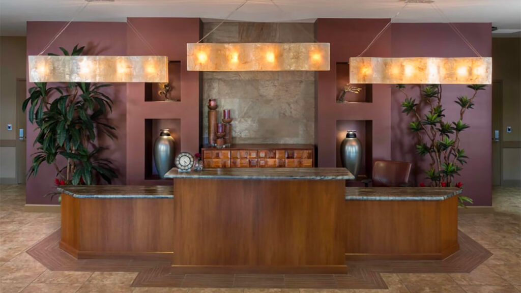 A modern reception desk with wood paneling sits between two large plants. Behind it are two decorative wall niches with vases, and above are two illuminated rectangular chandeliers. The walls are painted in warm tones.