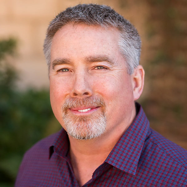 A smiling man with short gray hair and a goatee is wearing a dark red checkered shirt. He is standing outdoors with a blurred natural background.