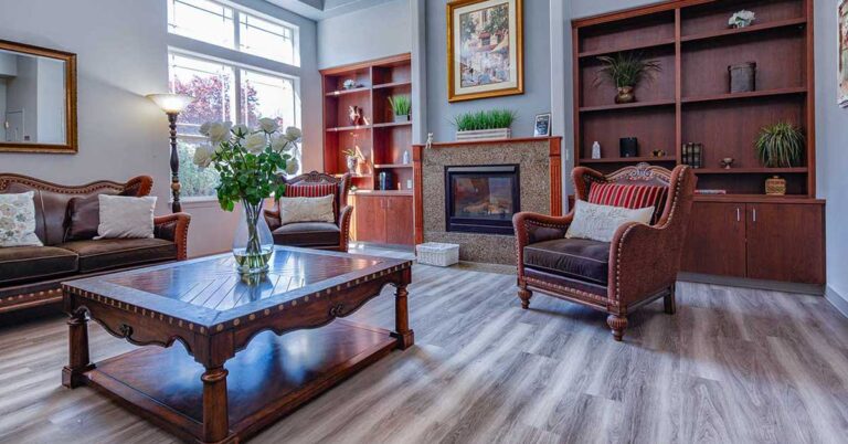 A cozy living room with a large wooden coffee table, surrounded by two sofas and an armchair. There's a fireplace between built-in bookshelves filled with decor. A floor lamp and a vase with white flowers add elegance. Natural light flows in through windows.