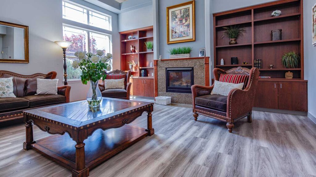 A cozy living room with wooden flooring features a fireplace, a framed art piece above it, and two elegant armchairs. There's a wooden coffee table with vases of flowers, flanked by built-in bookshelves with plants and decorative items.