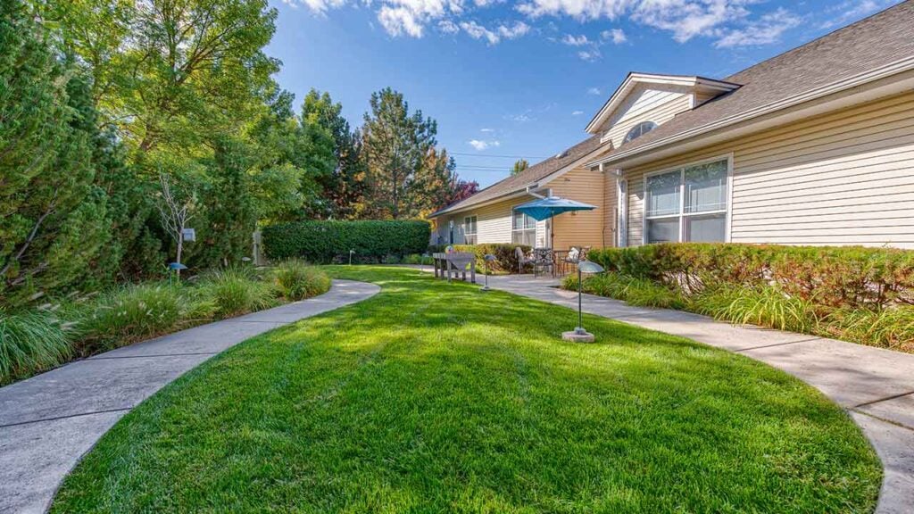 A well-maintained backyard with a lush green lawn, bordered by trees and shrubs. A curved concrete pathway leads to a seating area with a table and an umbrella, adjacent to a beige house with large windows. The sky is blue with wispy clouds.