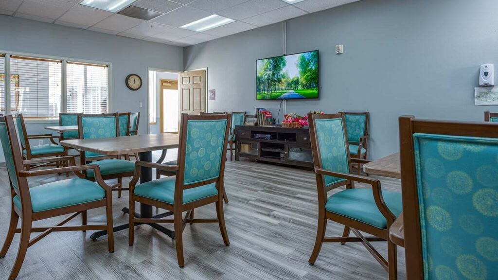 A spacious room with wooden tables and chairs with teal cushions. A large TV on the wall displays a nature scene. Light streams through a window, and the room has wood flooring and gray walls.