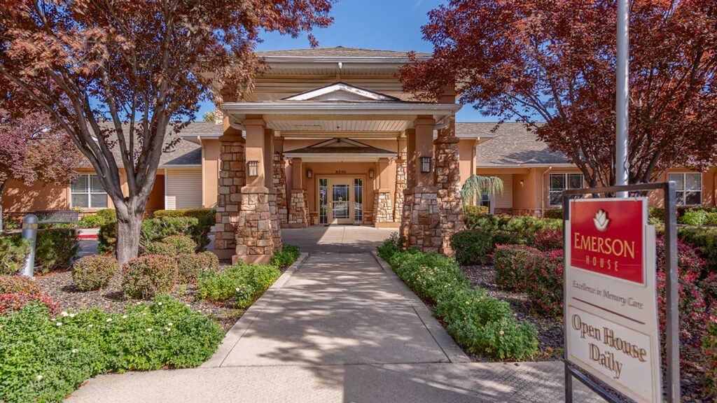 A welcoming entrance of a single-story building surrounded by lush landscaping and vibrant red-leaved trees. A sign in the foreground reads "Emerson House, Open House Daily.