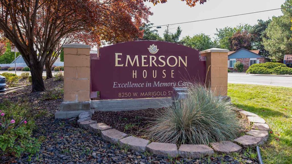 A sign that reads "Emerson House: Excellence in Memory Care, 8250 W. Marigold St." is surrounded by landscaping with a small garden and trees in the background.