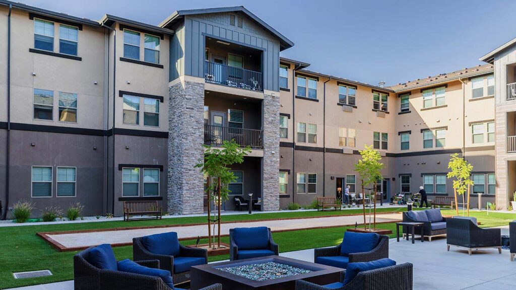 View of a modern apartment complex featuring a central courtyard with woven outdoor seating around a square fire pit. The building has three stories with balconies, and the landscaped area includes young trees and well-maintained grass.