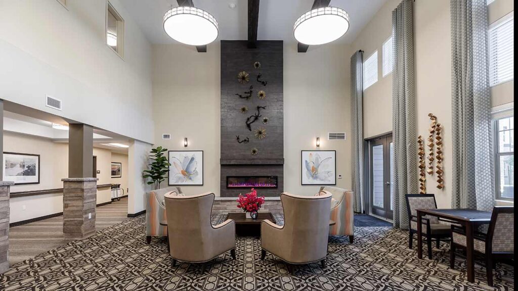 A modern living room with high ceilings, two beige armchairs facing a wall with abstract art and an electric fireplace. A table with chairs is on the right, and large circular light fixtures hang from the ceiling. Decorative elements adorn the walls.