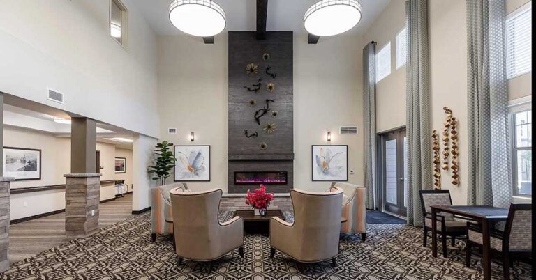 A spacious, modern lobby with high ceilings, featuring a central fireplace with decorative artwork above. There are elegant armchairs, a table with a plant centerpiece, large windows with curtains, and patterned rugs on the floor.