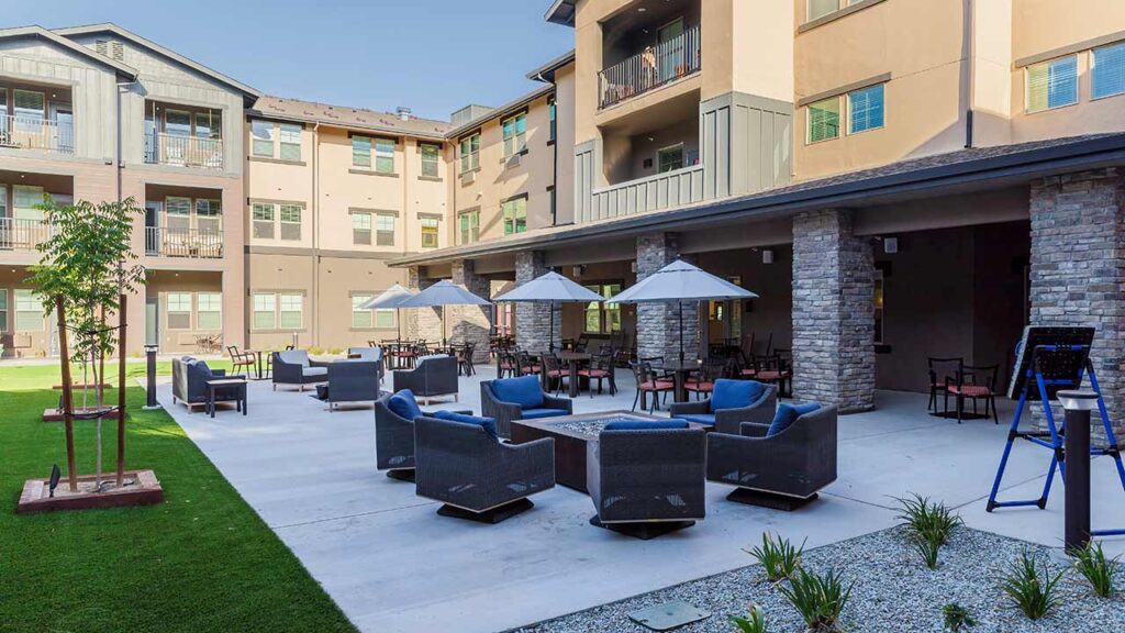 Outdoor patio area of a modern apartment complex featuring cushioned seating around a fire pit, tables with umbrellas, and landscaped greenery. The building has multiple stories with balconies overlooking the courtyard.