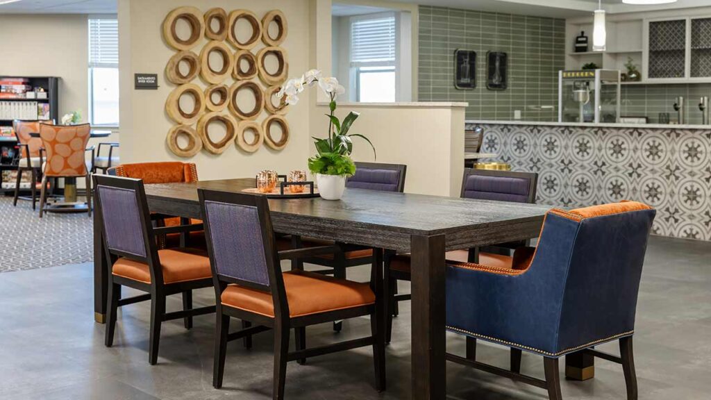 A modern dining area with a dark wooden table and six chairs, featuring orange and blue upholstery. The wall is decorated with circular wooden accents, and there's a countertop with intricate tile patterns in the background. A potted orchid sits on the table.