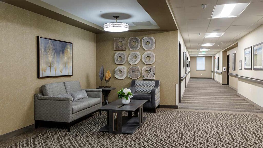 A waiting area with two cushioned chairs, a coffee table with flowers, artwork on the walls, and a carpeted floor. The area is well-lit, leading into a long corridor with more art and neutral-colored decor.