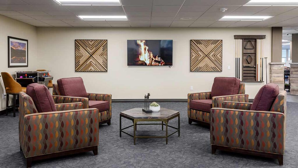 A cozy seating area with four patterned armchairs around a hexagonal coffee table. A wall-mounted TV displays a crackling fire. The room is decorated with geometric wall art and framed pictures. Neutral carpet and ceiling lights complete the space.